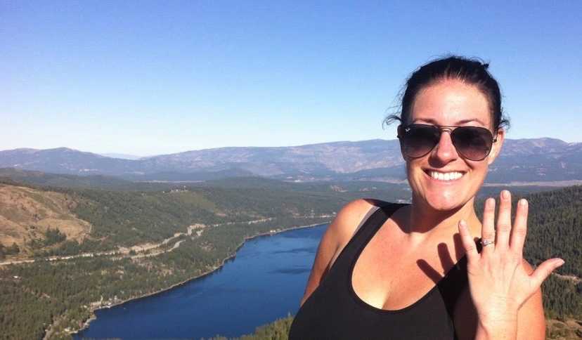 Woman smiles while holding up hand that has engagement ring on it with body of water in background