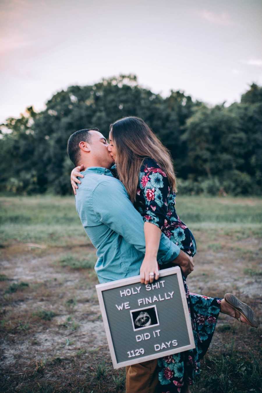 Husband holds up wife and kisses her as she holds sign that announced her pregnancy