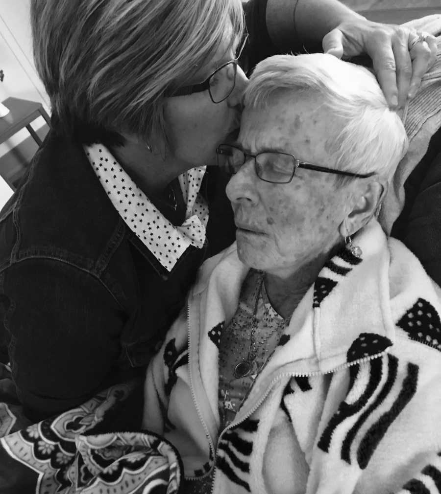 Elderly woman with dementia sits sleeping in chair while daughter kisses her forehead