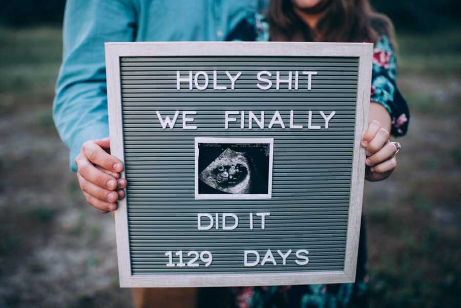 Husband and wife hold up sign that says, "Holy Shit We Finally Did It 1129 Days" with sonogram