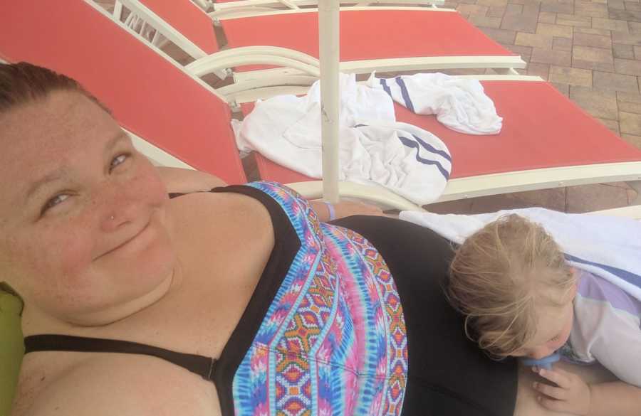 Plus sized mom wearing a swimsuit smiles in pool chair with child lying on her lap