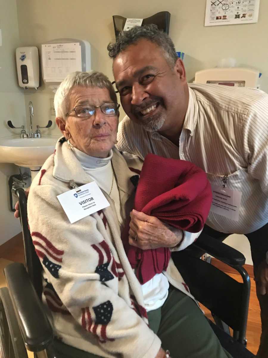 Woman with dementia sits in wheelchair in nursing home with man smiling beside her