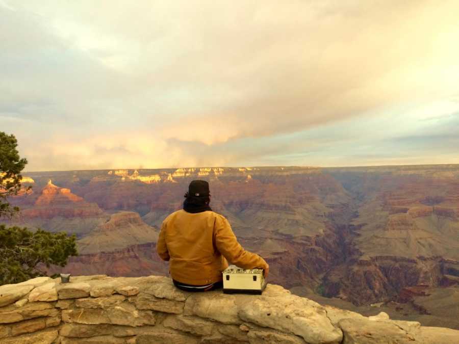 Man sits on stone wall with hand on wife's ashes looking out at Grand Canyon