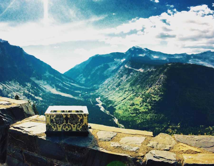 Box of woman's ashes sitting on stone wall with mountains in background