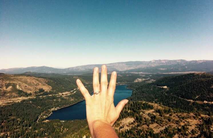 Hand with engagement ring on it is held in air with mountains and body of water in background