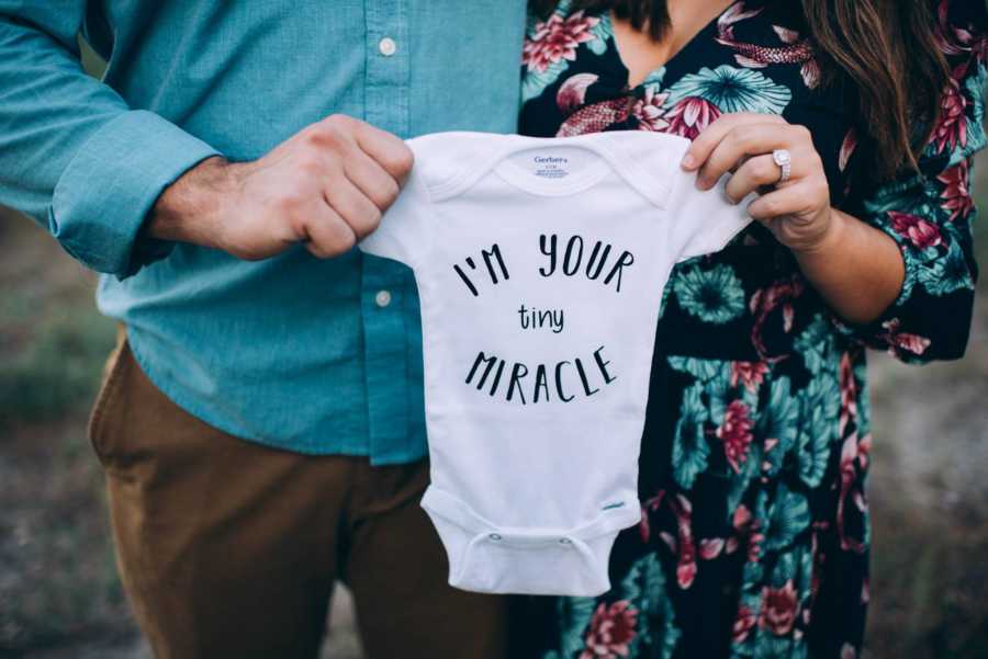 Husband and wife hold up onesie that says, "I'm your tiny miracle"