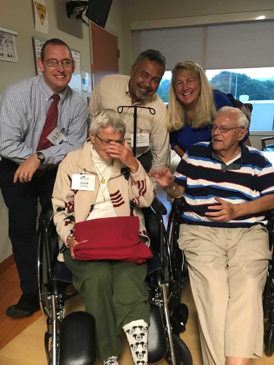 Daughter stands behind father and mother with dementia sitting in wheelchairs beside two men