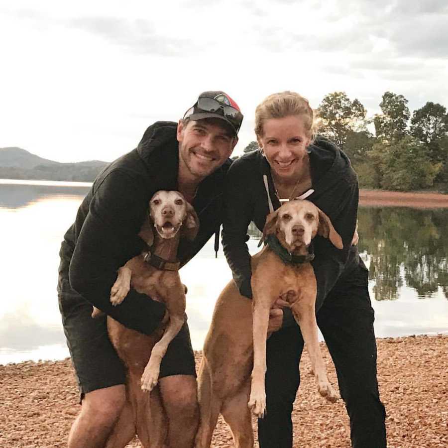 Husband and wife who struggle to get pregnant stand on shore of water holding onto their two dogs