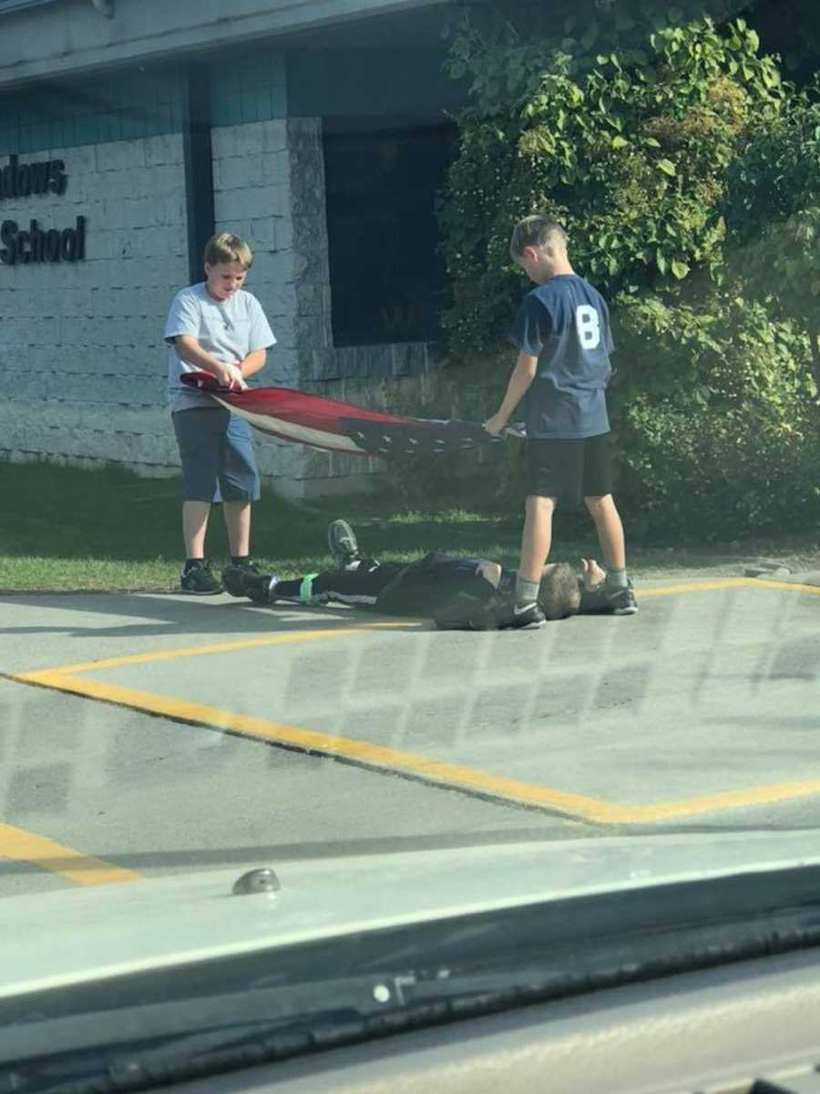 Two boys holding American Flag in parking lot while another boy lays under it so it won't touch the ground