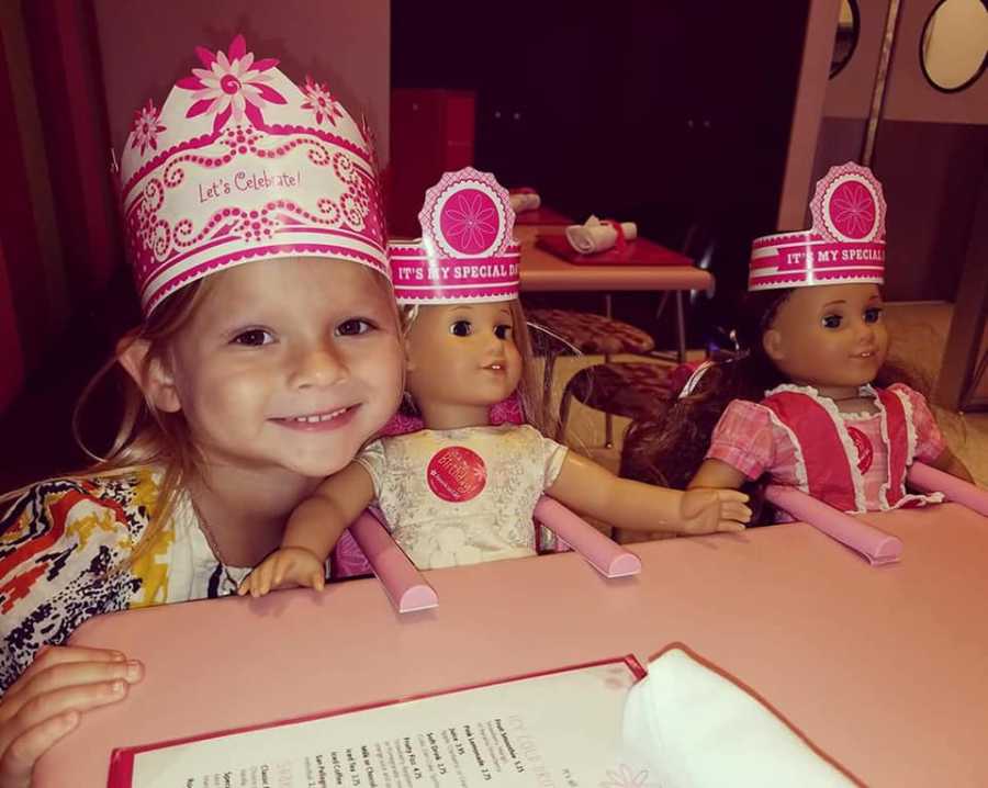 Little girl who has since passed from sepsis smiles at restaurant table beside two American Girl Dolls