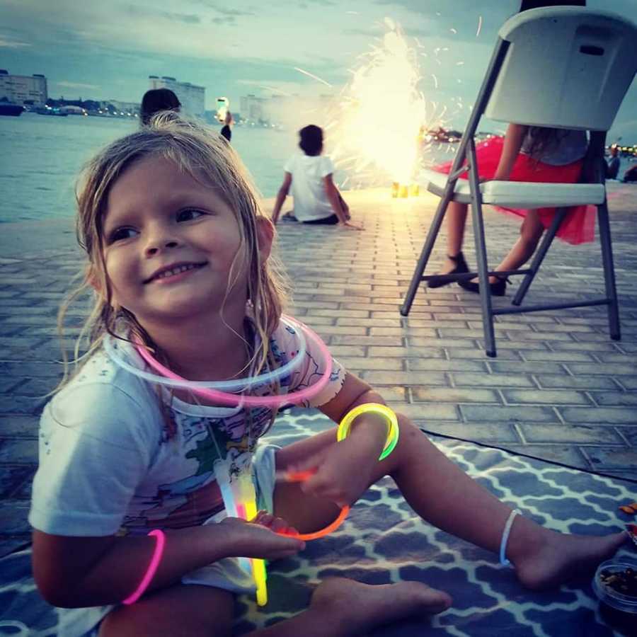 Little girl with sepsis smiles with glow stick jewelry on in front of body of water and fire works