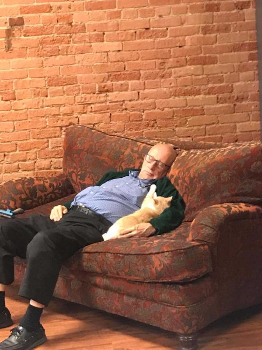Man asleep on couch of cat shelter with cat lying in his arm