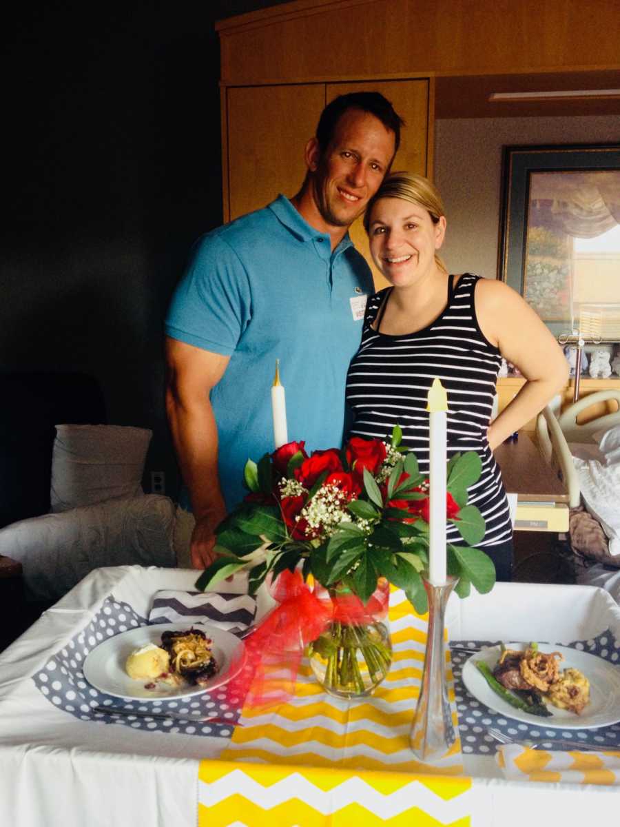 Husband and wife stand smiling behind set dinner table in hospital room