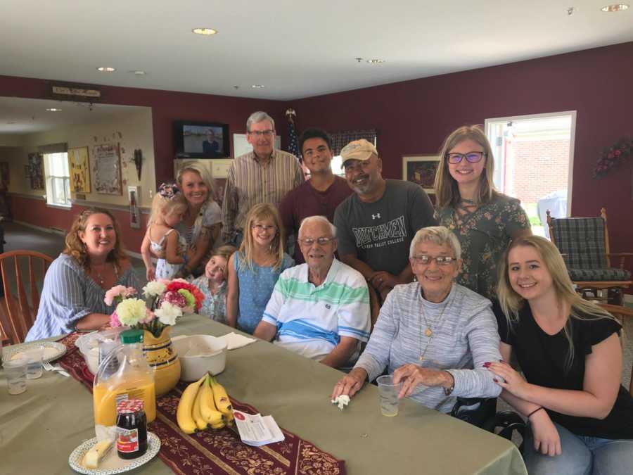 Woman with dementia sits at table in nursing home beside husband and family members