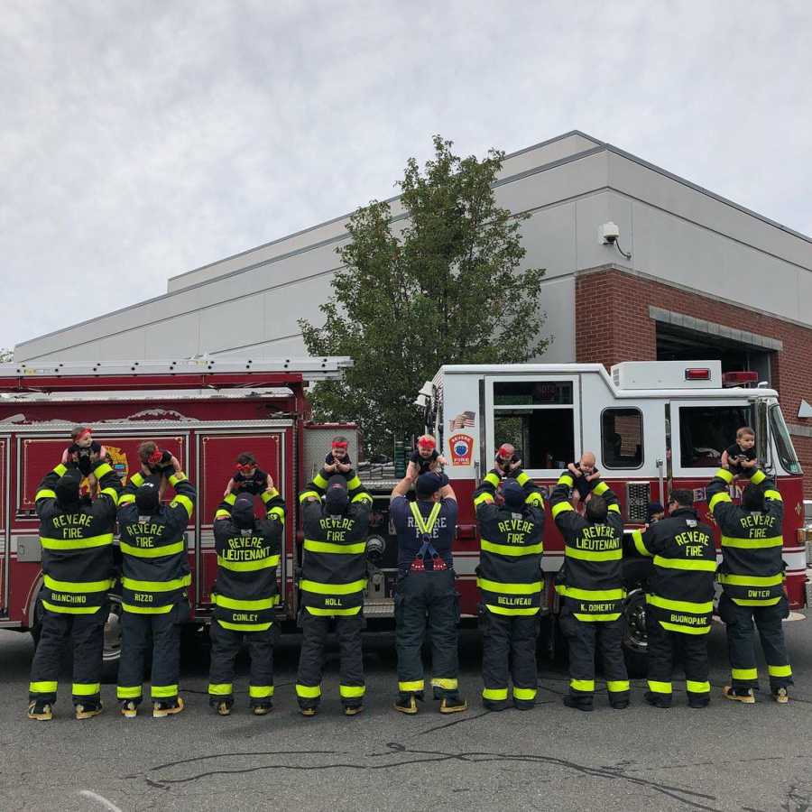 Nine firefighter stand in front of fire truck holding their newborn babies up in the air