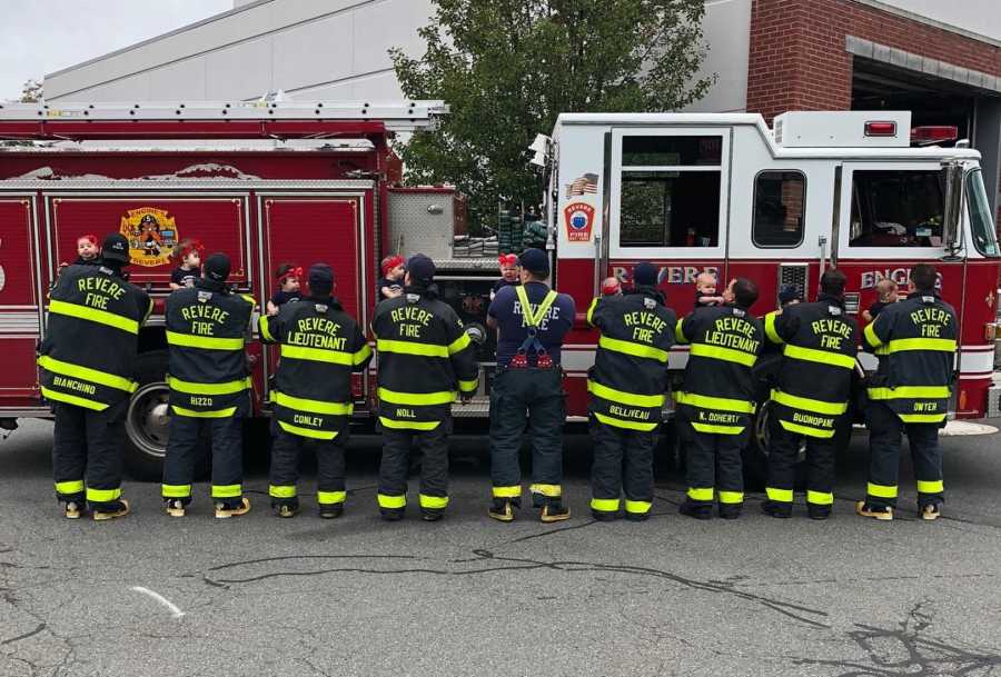 Nine firefighters stand facing fire truck holding their newborns 
