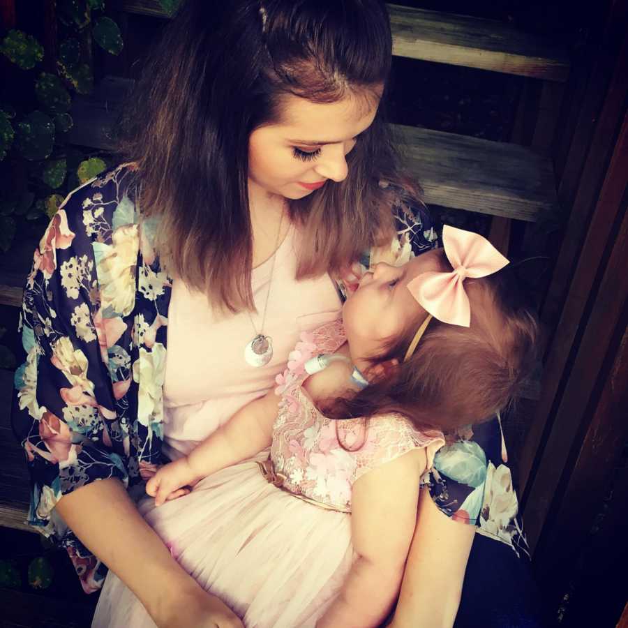 Mother looks down at toddler with Trisomy 18 sitting in her lap in pink dress and head band with bow