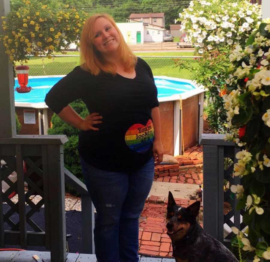 Pregnant woman who lost her first child stands with hand on her hip on back porch beside dog 