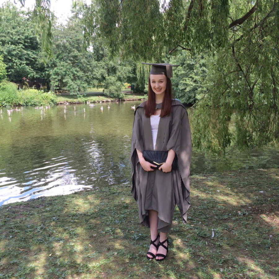 College graduate with M.E. stands smiling in cape and gown near pond