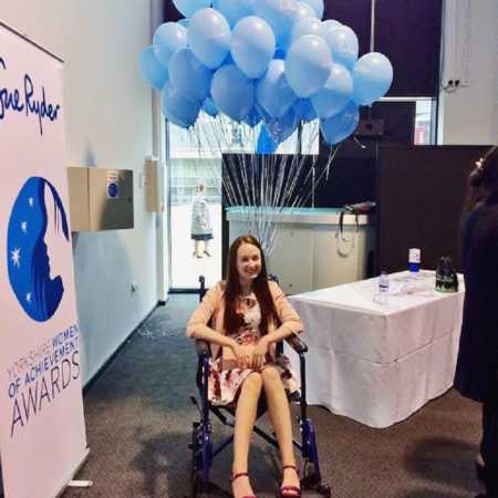 Young woman with disability smiles in wheelchair with blue balloons attached beside sign for an award ceremony