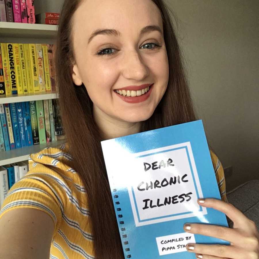 Young woman with disability smiles in selfie holding up book she wrote called, "Dear Chronic Illness"