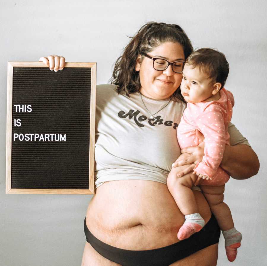 Woman with shirt pulled up to show her stomach holds daughter and sign saying, "this is postpartum"