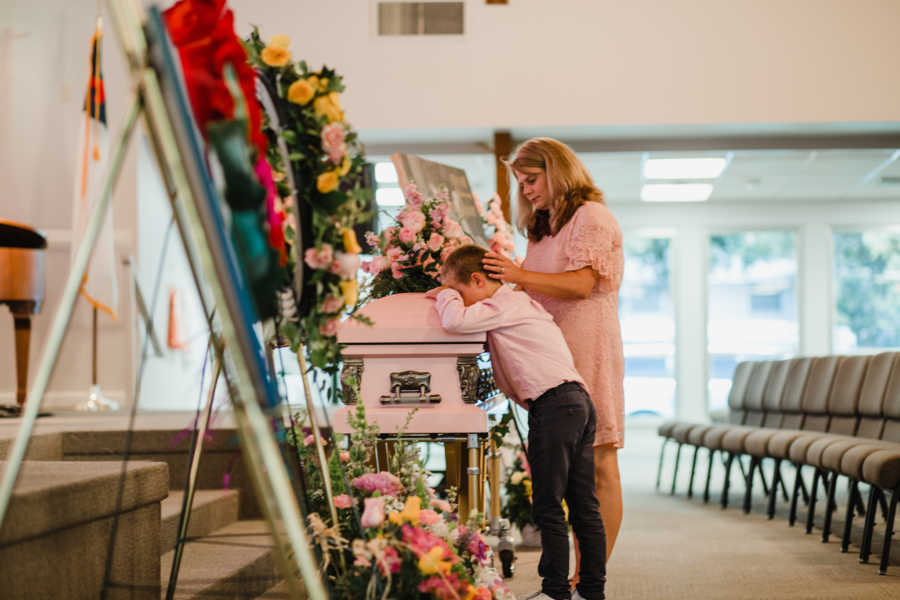 Little boy rests his head on coffin crying while woman stands beside him with her hand on his head