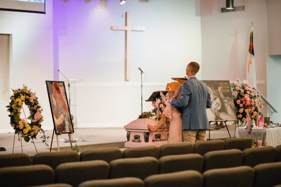 Husband stands behind wife holding her at daughter with sepsis funeral as they look up at large cross