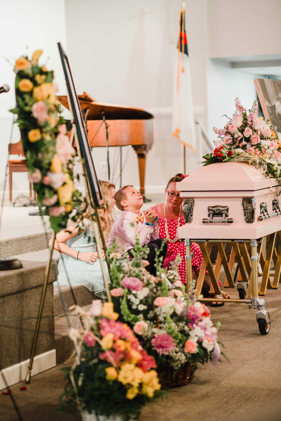 Little boy sits next to little girl with sepsis' coffin holding his hands together crying 