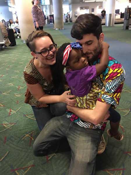 Husband and wife kneel on floor of airport while husband holds adopted daughter in his arms