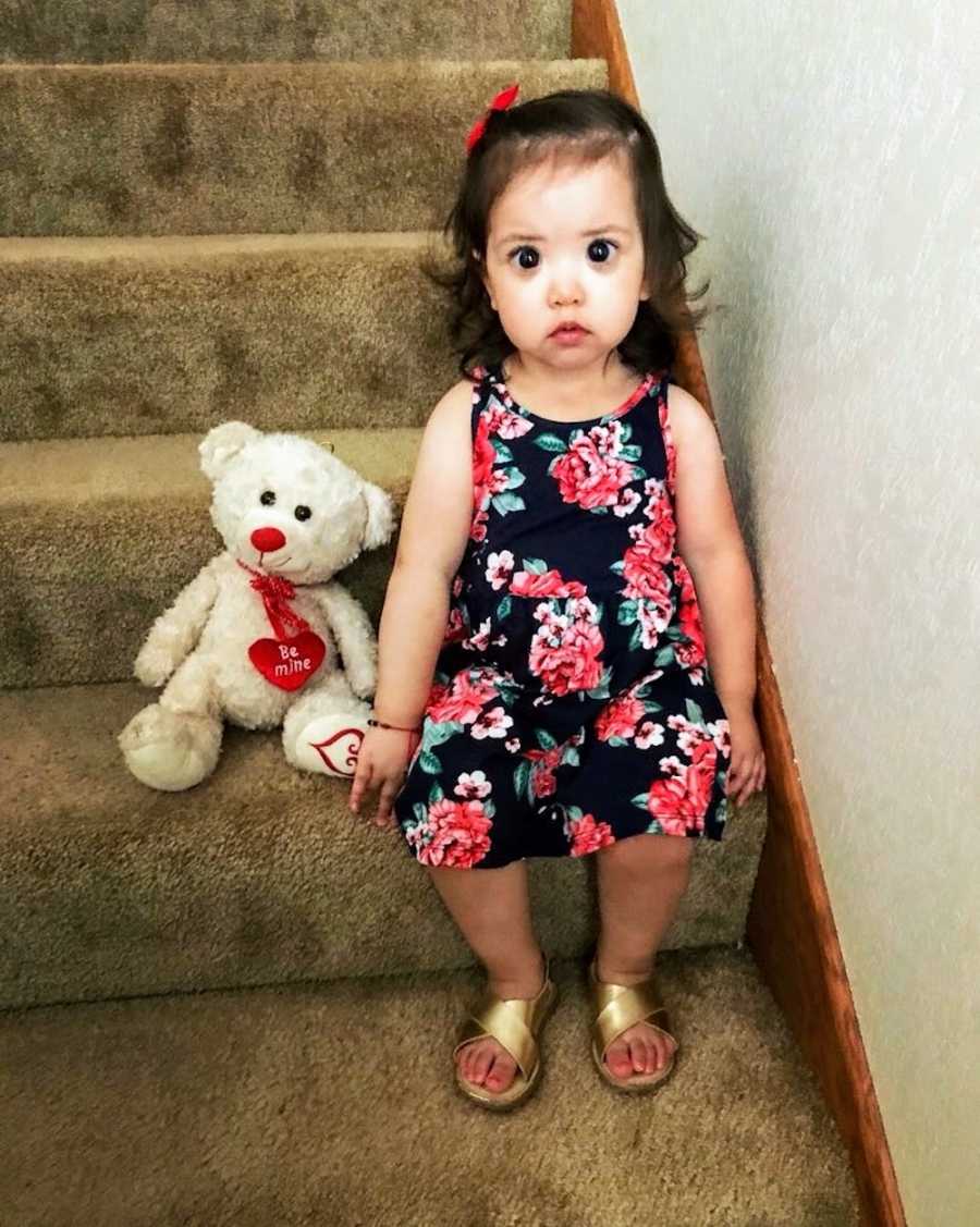 Toddler with Axenfeld-Rieger syndrome sits on staircase in floral dress beside teddy bear