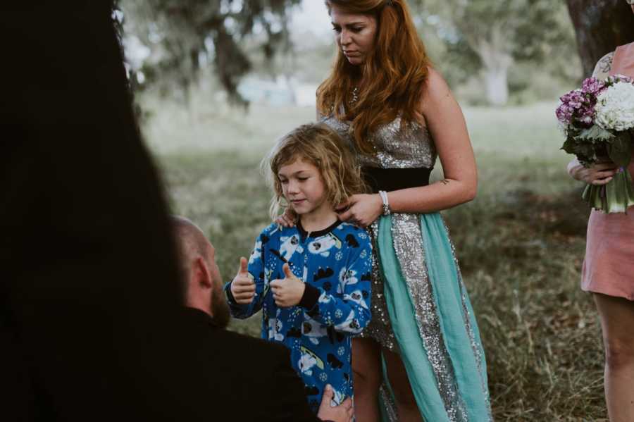 Mother stands behind son who is holding two thumbs up in front of mothers new husband