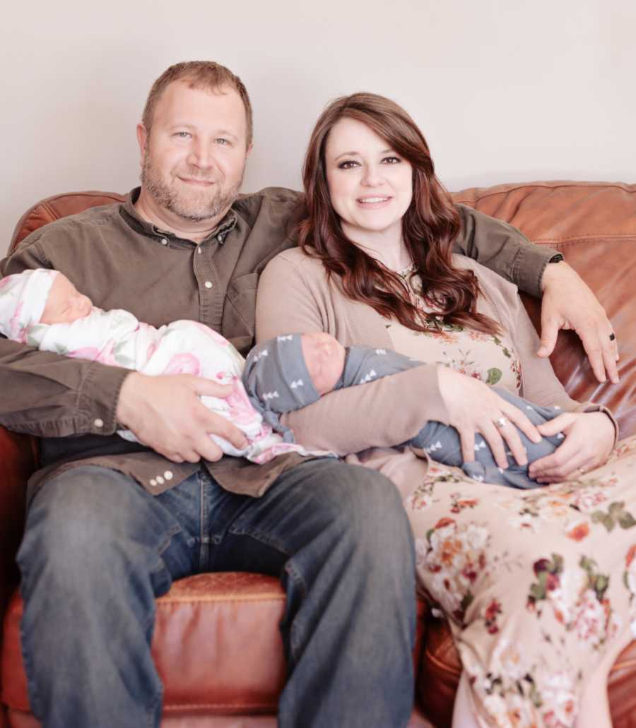 Husband and wife sit smiling on the couch with their newborn twins in their lap
