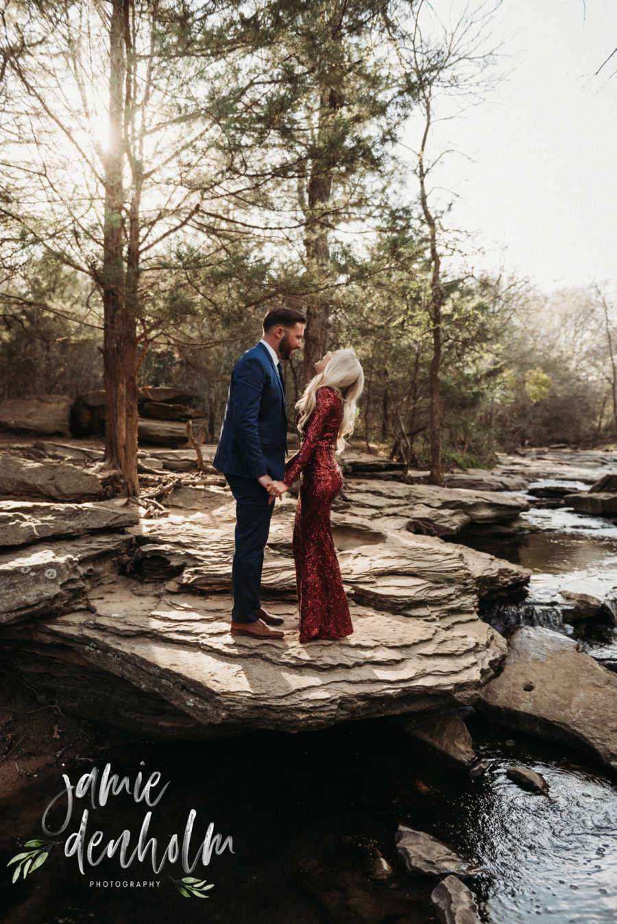 Husband and wife stand holding hands in formal attire on rock beside river