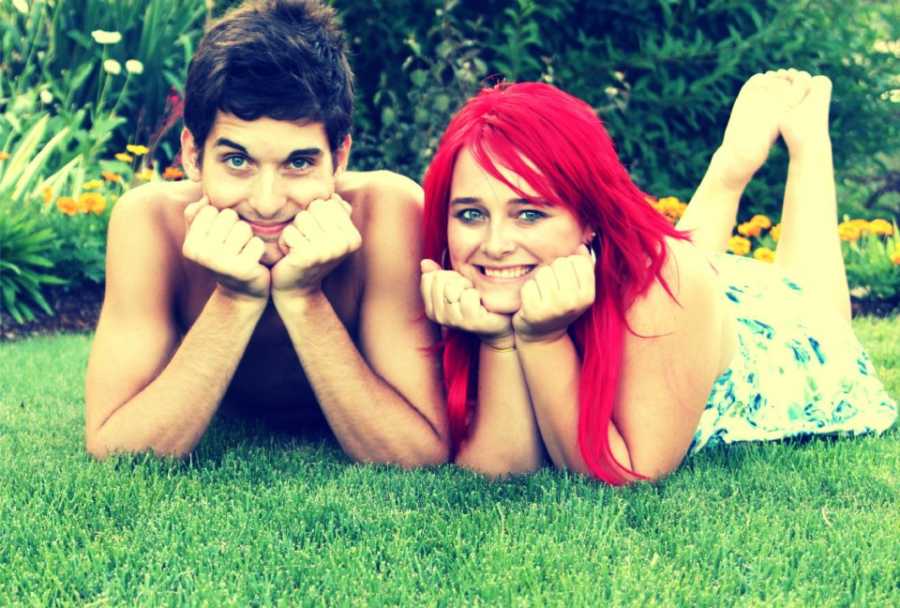 Boyfriend and girlfriend lay on grass with their chins resting on their hands smiling