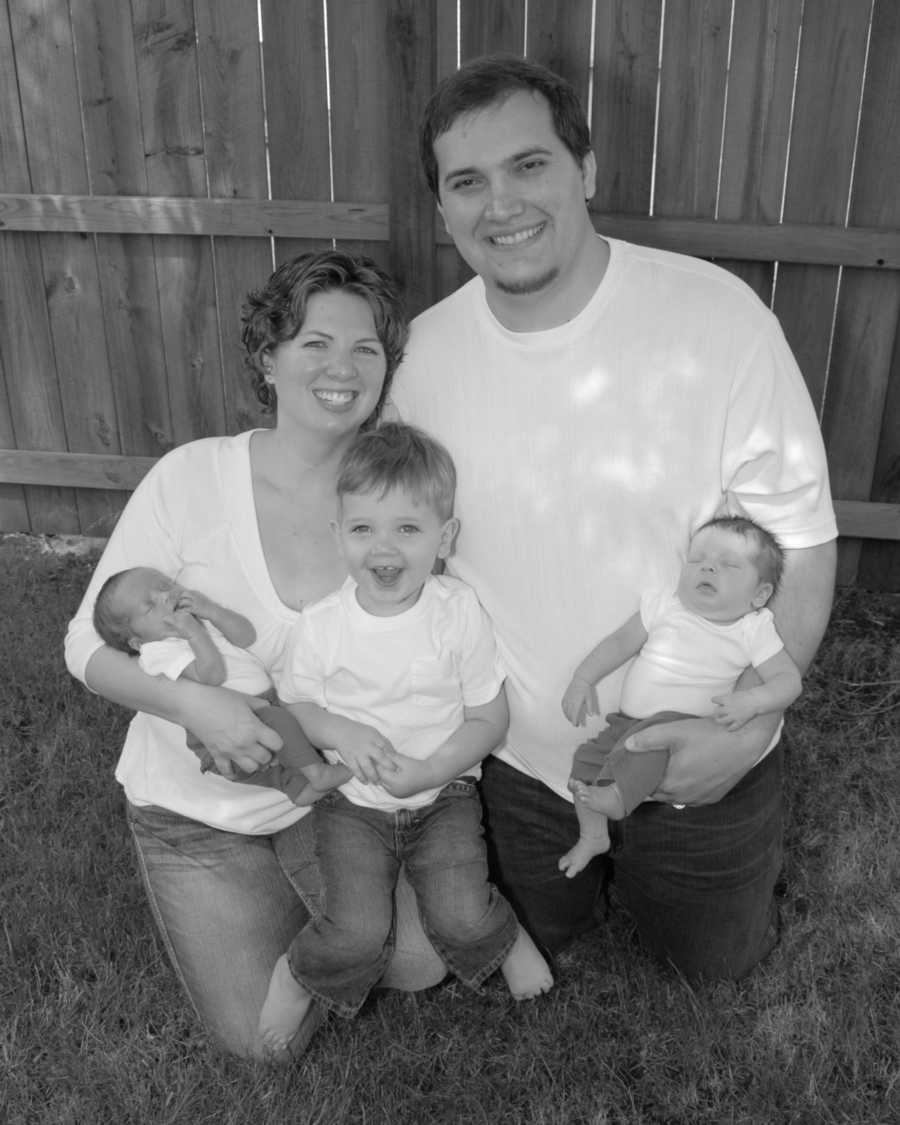 Mother who had Hodgkin Lymphoma and hysterectomy kneels on ground with two sons in her lap beside husband and other son