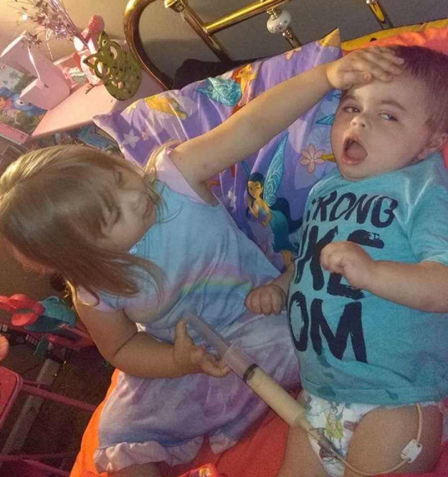 Older sister sits in bed holding hand on little brothers forehead who had a brain injury