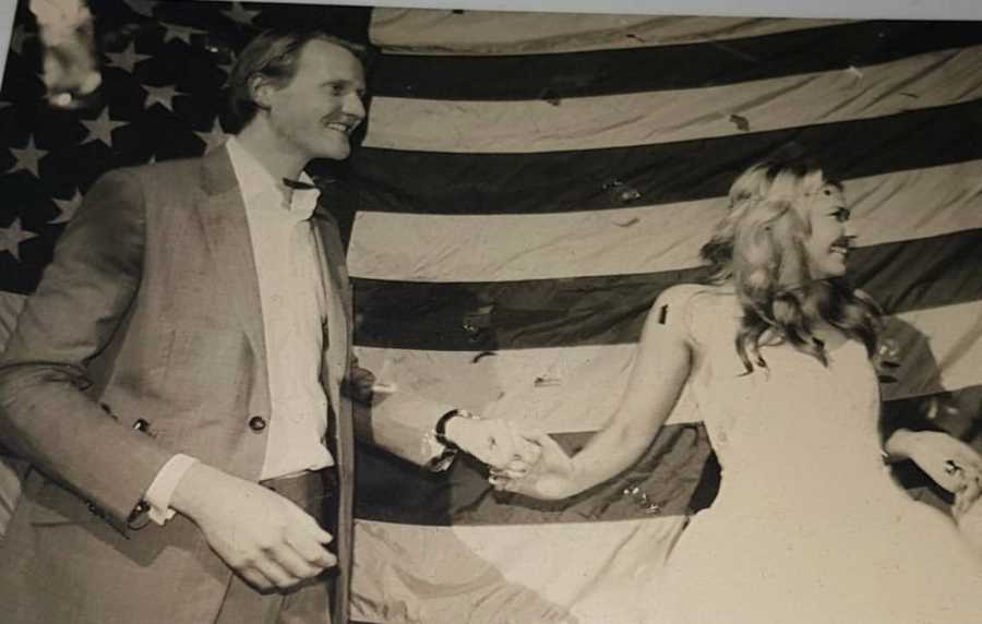 Husband and wife holding hands in front of large American Flag