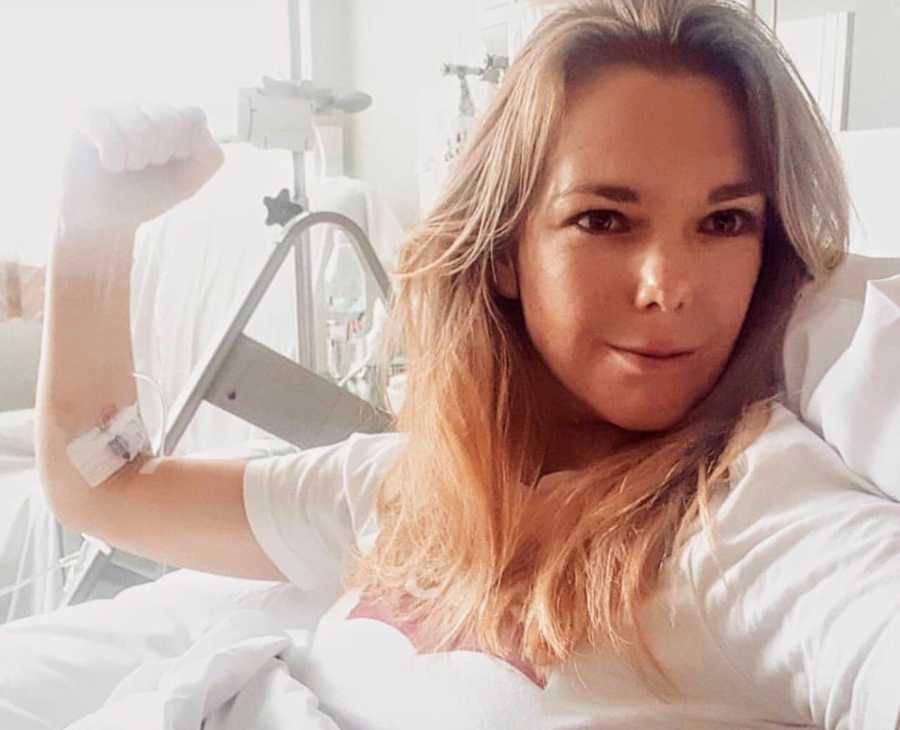 Woman who struggles with fertility sits in hospital bed flexing arm with IV in it in selfie