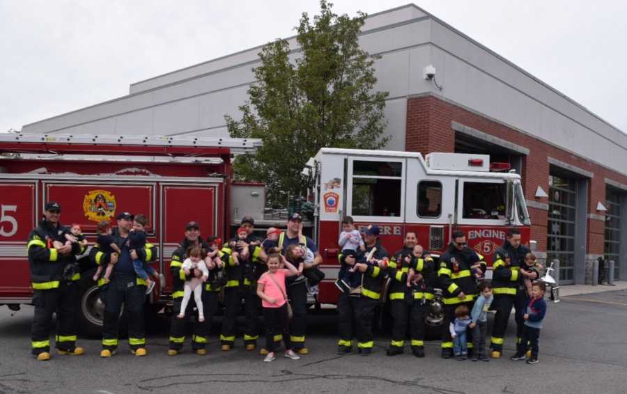 Nine firefighters stand in front of fire truck holding their newborns and their other children 