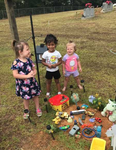 Three little girls stand beside grave of baby who passed away with toys surrounding it