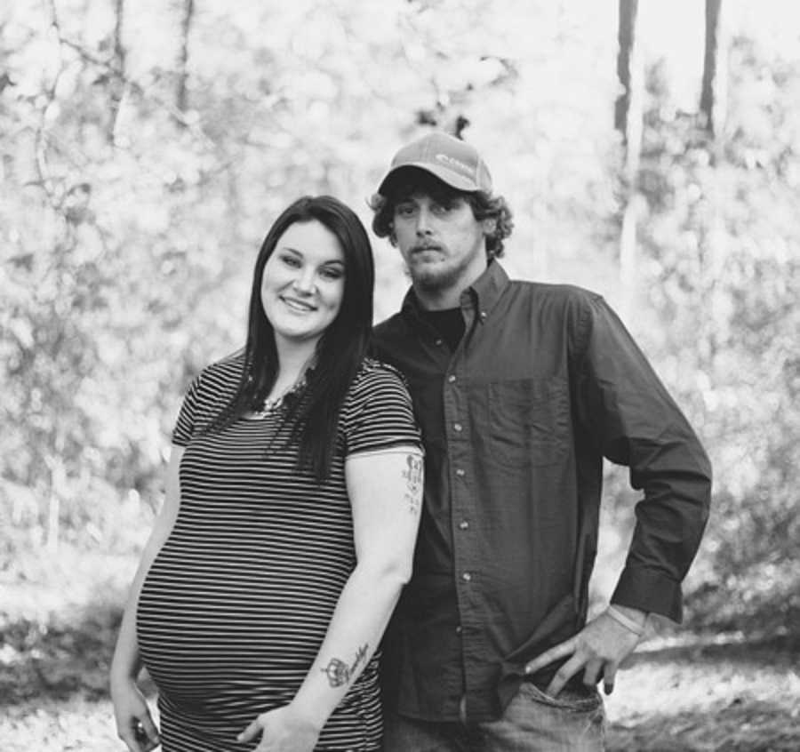 Pregnant wife stands smiling beside husband who stands with hand on his hip