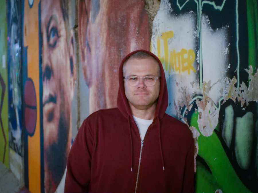 Man who was once a junkie and is now an activist stands against graffiti wall with hood up