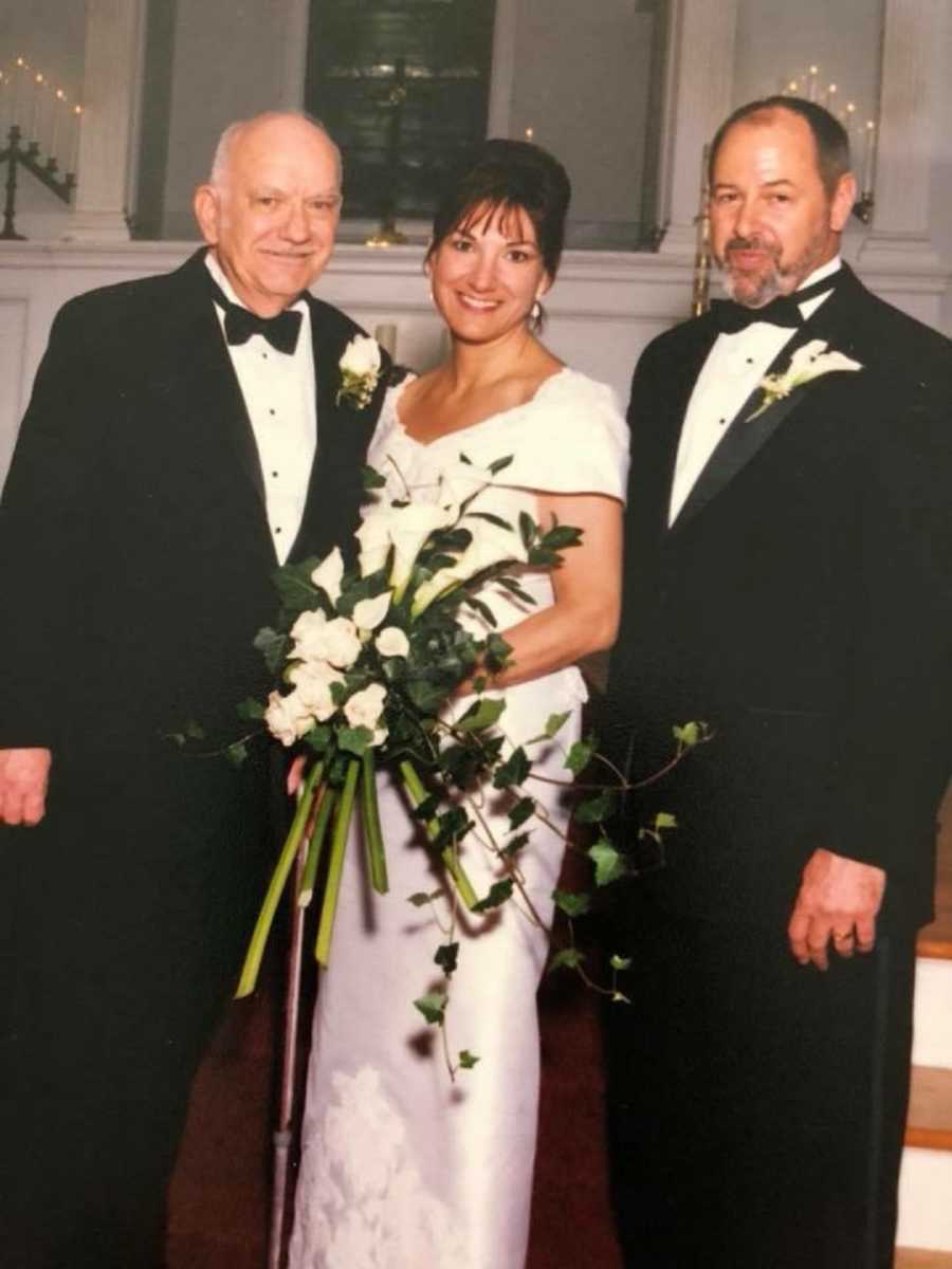 Bride smiles beside groom and father while holding bouquet of flowers