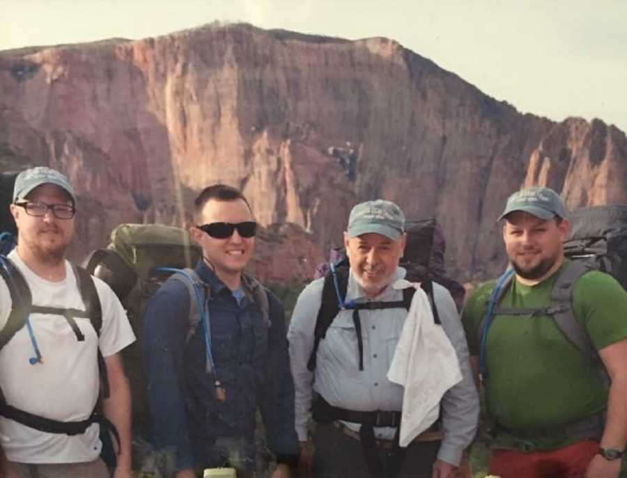 Man with stage 4 cancer stands beside three sons with hiking backpacks on and mountain in background