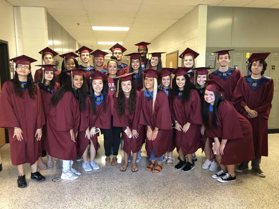 Group of high school graduates in cap and gown who has inspired their teacher who is standing amongst them