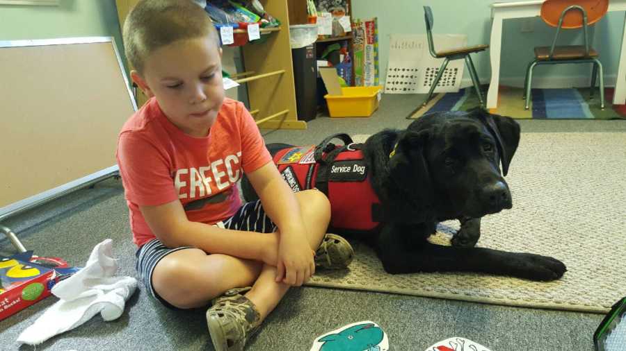 Little boy with autism and speech apraxia sits on floor in classroom beside his service dog