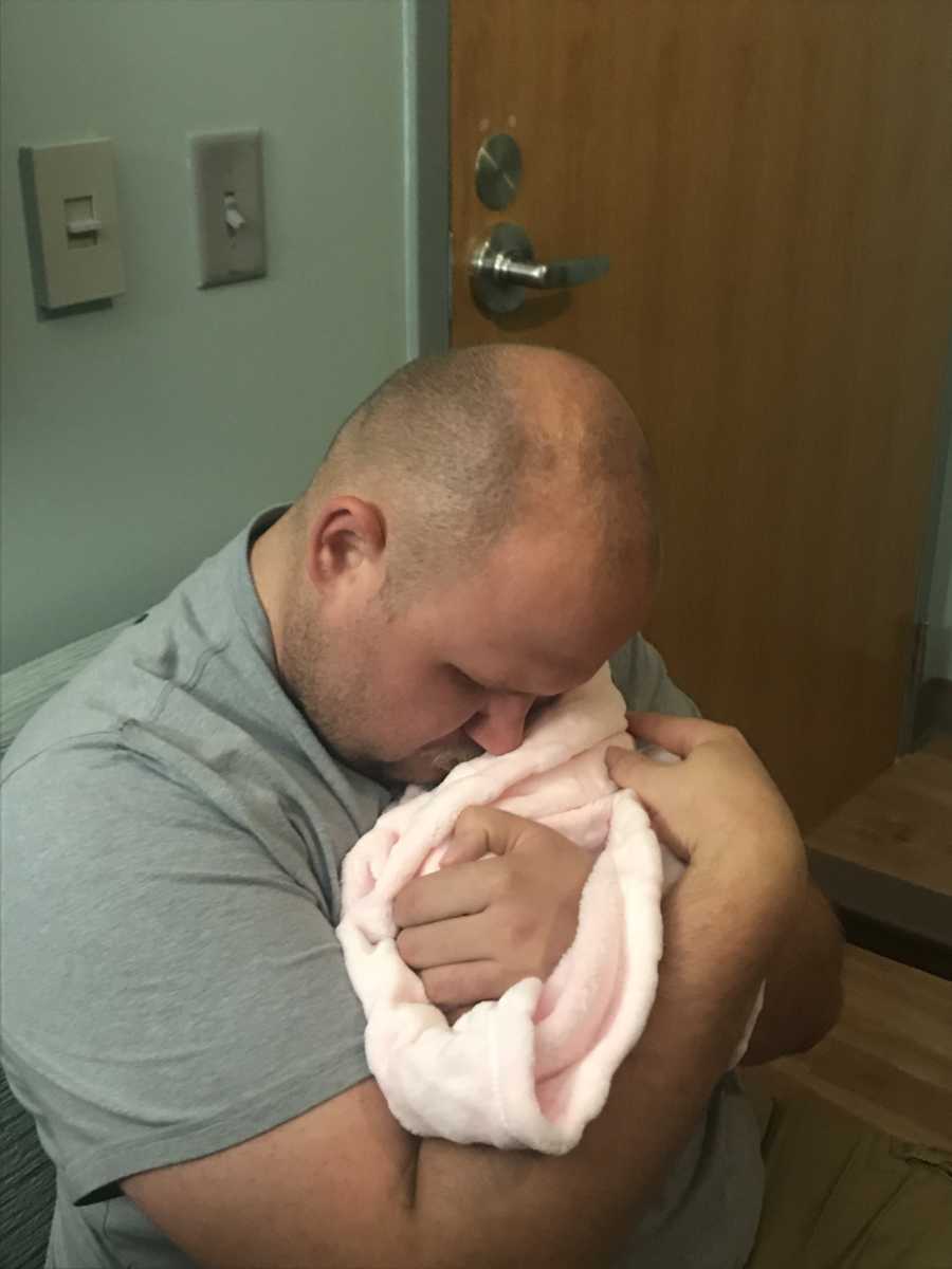Father sits holding micro preemie in pink blanket with his arms wrapped around her