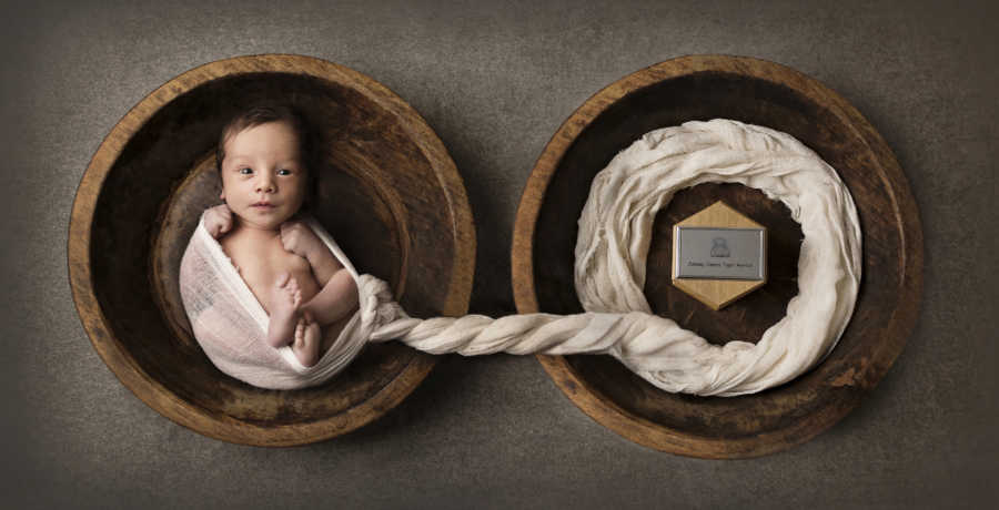 Newborn sits on bowl wrapped in cloth that is connected to another bowl with plaque of deceased twins name