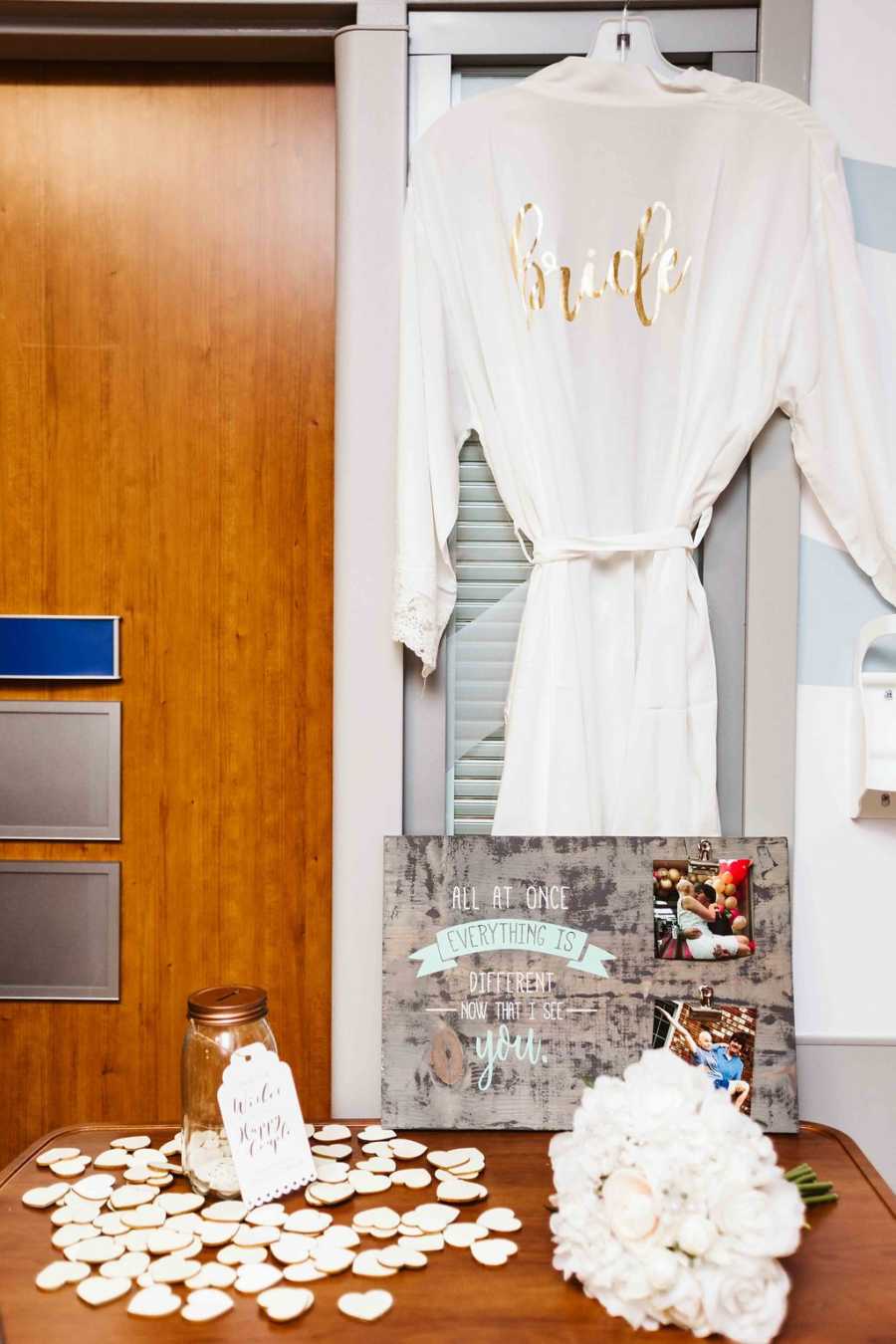 White robe hanging in hospital room that says, "Bride" in gold behind next to table with flowers and candle